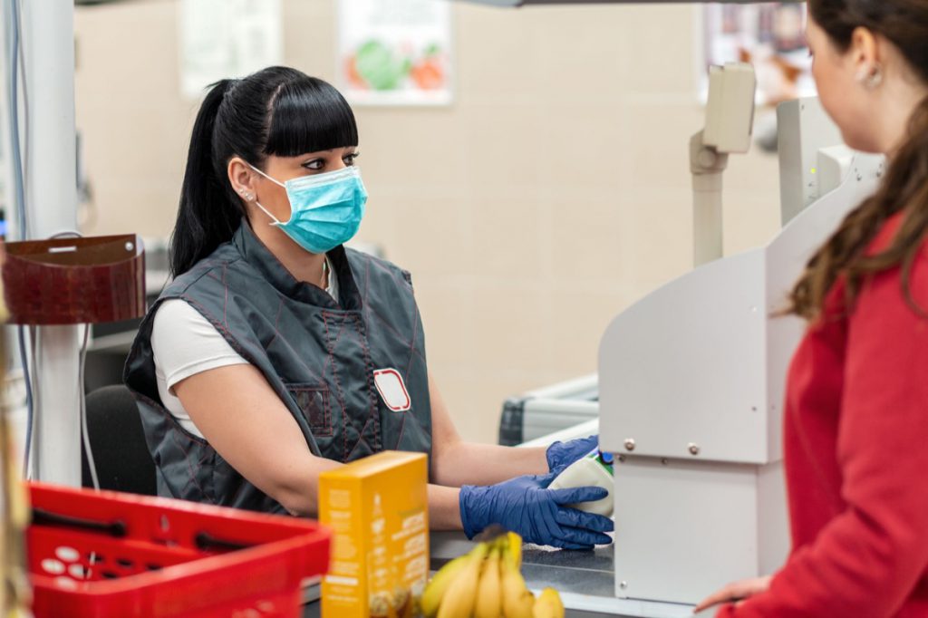 A young woman in a medical mask and gloves, working at the checkout in a supermarket. In the foreground is a customer in a blur. Close up. Concept of coronovirus, protection from infection and industrial crisis.