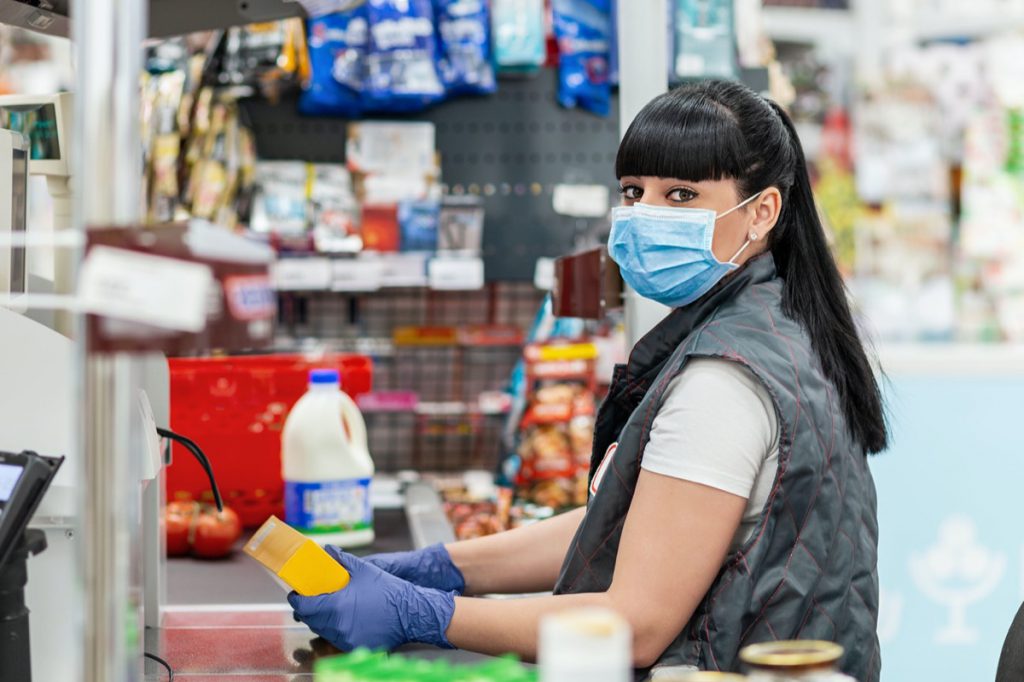 A portrait of young woman in a medical mask and gloves, working at the checkout in a supermarket. Concept of coronovirus, protection from infection and industrial crisis.
