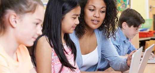 Teacher Helping Group Of Elementary School Children In Computer Class