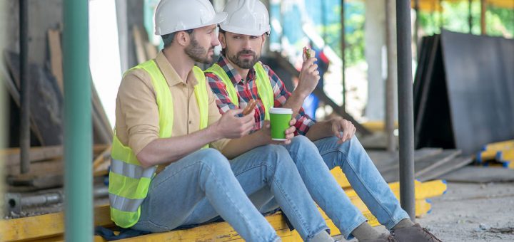 Building site. Building workers in yellow vests and helmets sitting on boards, having coffee with sandwiches, discussing something