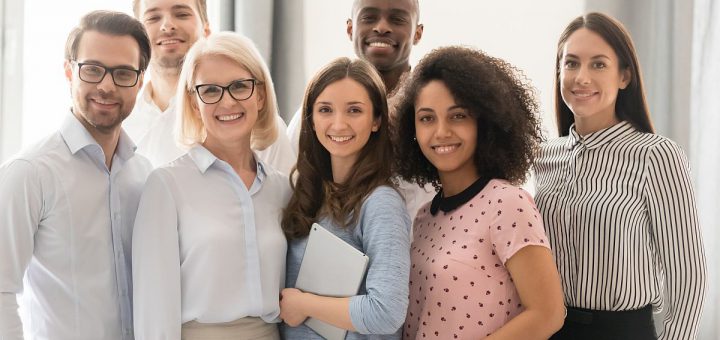 Multiethnic smiling businesspeople standing looking at camera making group photo in office together, happy diverse employees posing for picture with boss or team leader, showing unity and support