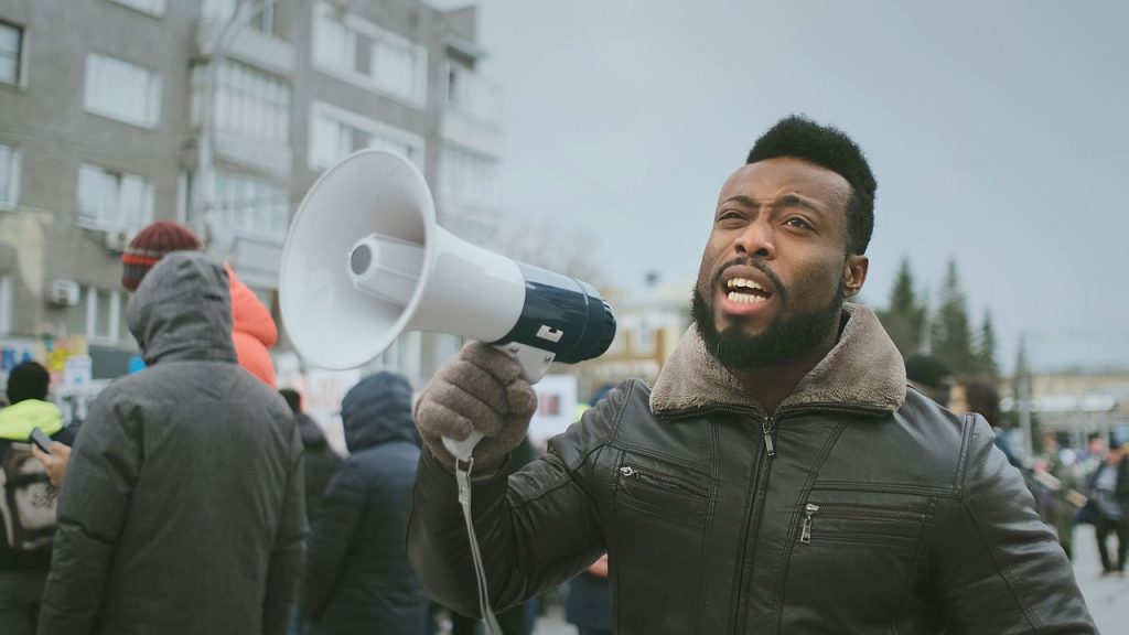 African american man hold megaphone hand and shouting. Crowd people day demonstration. Afro rebel strike protest. Revolution city street. Political rally. Loudspeaker shout. Activists outdoor fight.