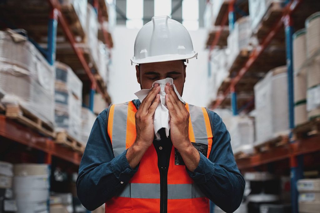 Worker wearing a hardhat in a warehouse blowing his nose; paid sick leave concept