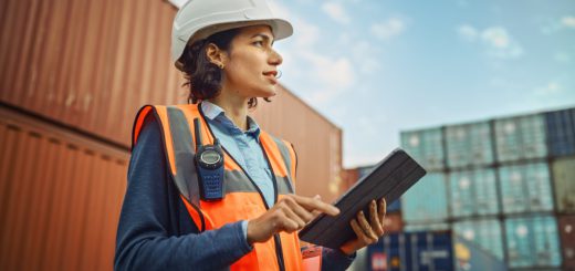 Worker in white hard hat and high-visibility vest working on tablet in container terminal; Department of Labor initiatives concept
